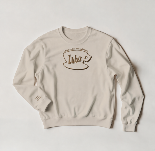 A light beige crewneck sweatshirt with a brown graphic on the chest that reads 'I drink coffee like a Gilmore' above a coffee cup outline containing the name 'Luke's'.