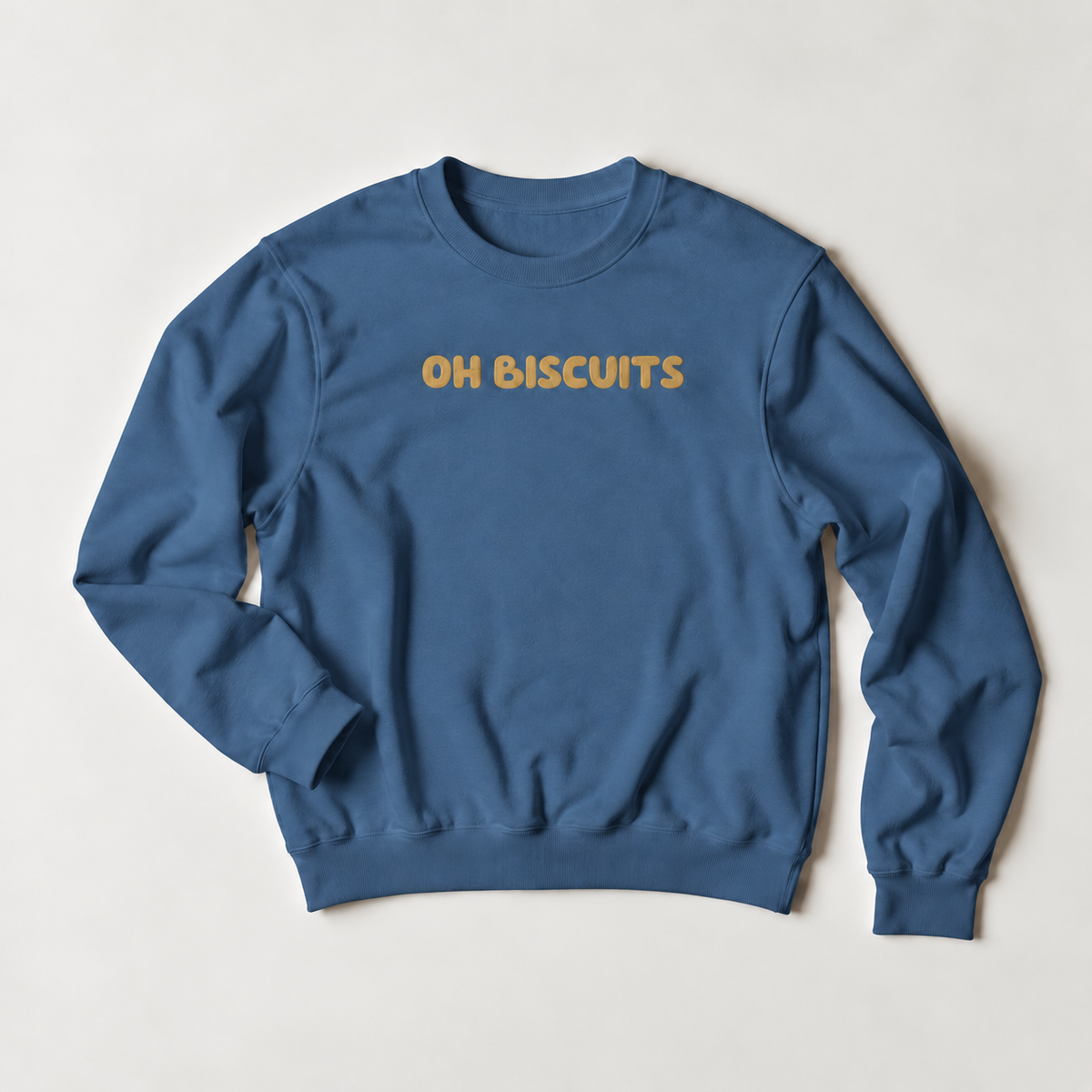 A blue crewneck sweatshirt with the words 'OH BISCUITS' printed in a warm yellow-orange font across the chest, laid flat on a light beige background.