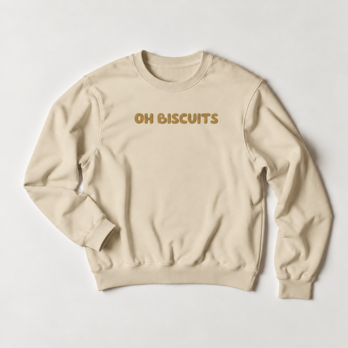 A beige crewneck sweatshirt with the phrase 'OH BISCUITS' printed across the chest in a textured mustard-gold font, laid flat on a light gray background.